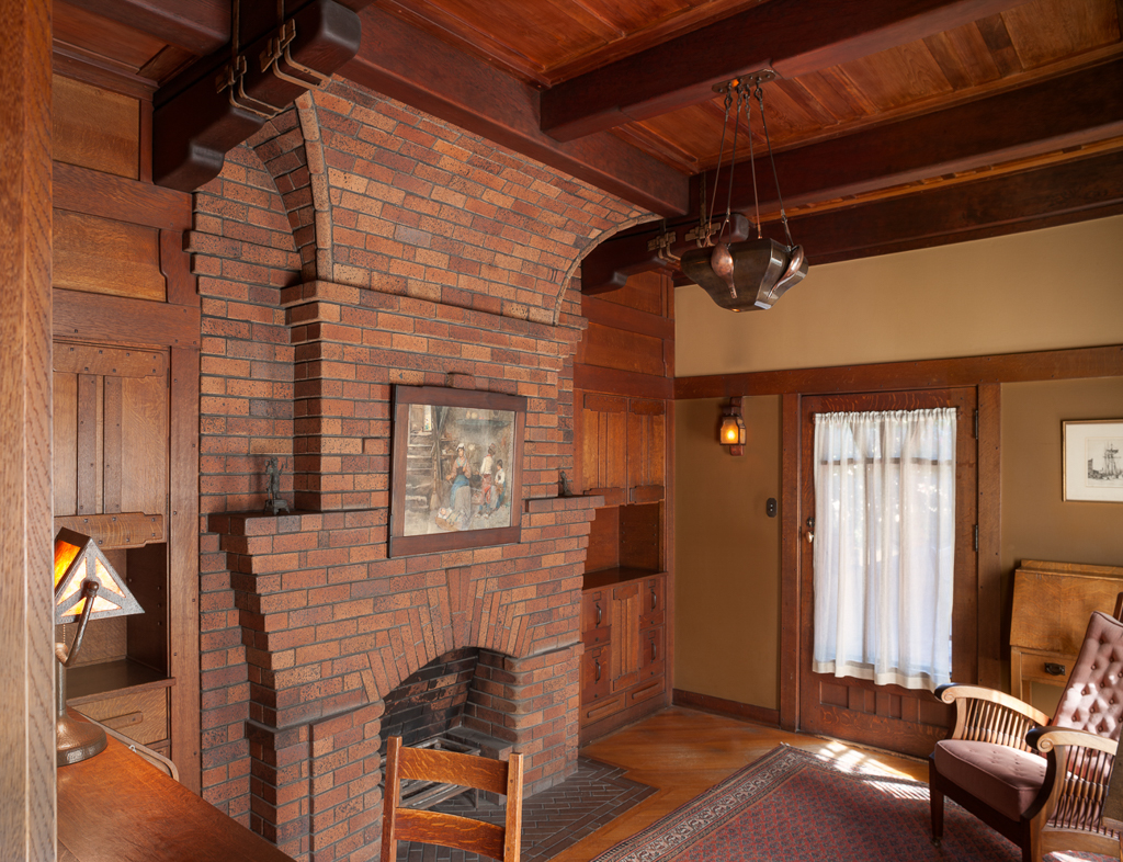 Interior photograph of the den at the Gamble House by Greene and Greene, showing a large red-brick fireplace with an arched firebox, brick chimney breast rising into a beamed wood ceiling, with built-in oak cabinetry, a hanging mica lantern, and Arts and Crafts furnishings