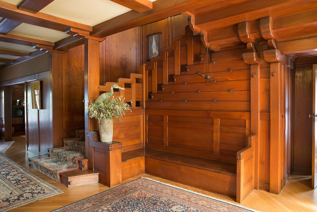Interior view of the David B. Gamble House entry hall showing the teak staircase, built-in bench, expressed structural beams with cloud-lift profiles, ebony peg details throughout the paneling, and an Asian-influenced bronze hanging sculpture