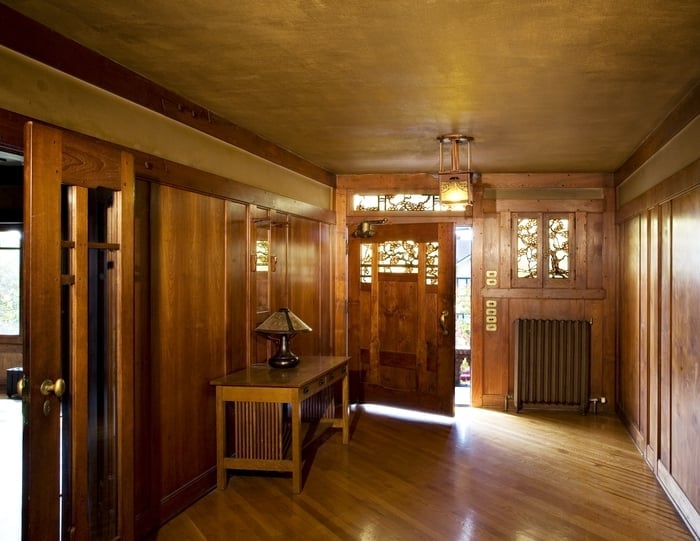 The Thorsen House entrance hall seen from inside, the mahogany-paneled door with its glowing art glass, a copper lantern above, and stacked brass push-plates beside the door.
