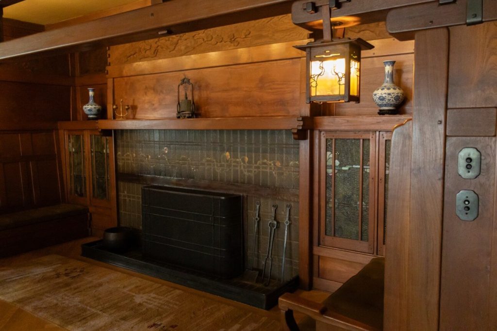 Interior photograph of a Greene and Greene inglenook with a low fireplace framed in iridescent green Grueby tile, flanked by quarter-sawn oak built-in cabinets with leaded art-glass doors, a hanging copper-and-mica lantern, and a carved wood overmantel