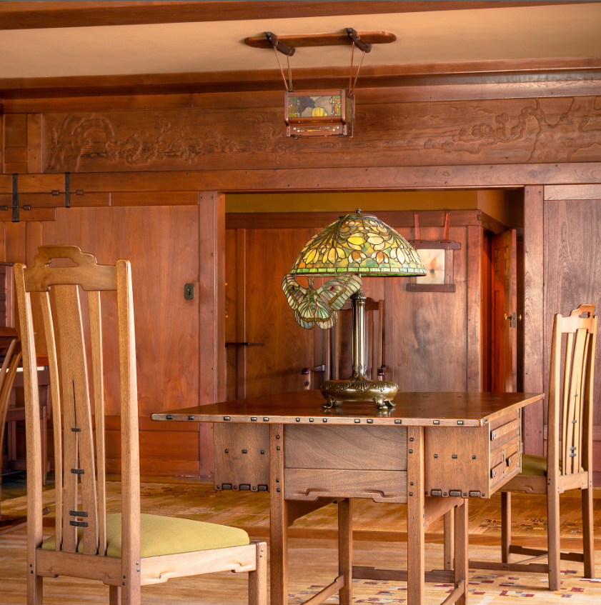 Detail of the original Greene and Greene library table in the David B. Gamble House, showing the cloud-lift apron, expressed ebony pegs, and tapered legs, with a Tiffany butterfly art glass lamp on top and Greene-designed dining chairs alongside