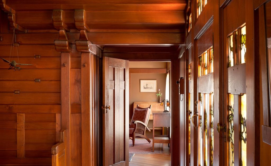 Interior view through a doorway of the David B. Gamble House, showing the layered teak paneling, expressed structural beams with rounded brackets, art glass panels filtering warm light from the entry hall, and a glimpse of an adjoining office with a craftsman desk and chair
