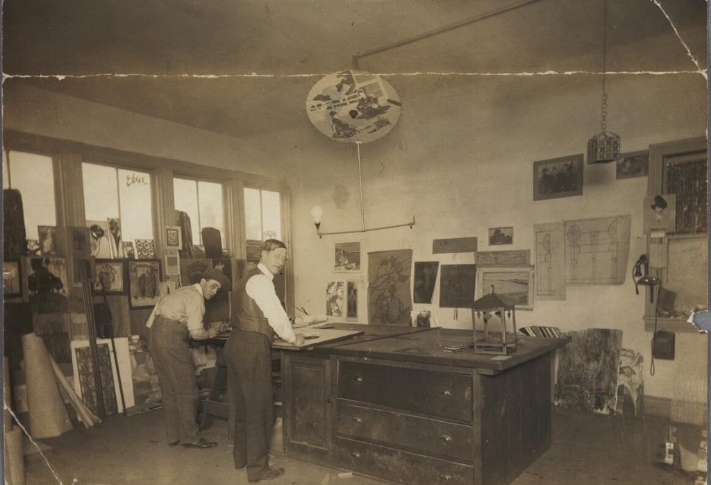 Sepia historic photograph of Emil Lange and a colleague working at drawing boards in the Lange art glass studio in Los Angeles, showing completed art glass panels on the walls, design sketches, and architectural drawings of leaded glass windows, early twentieth century