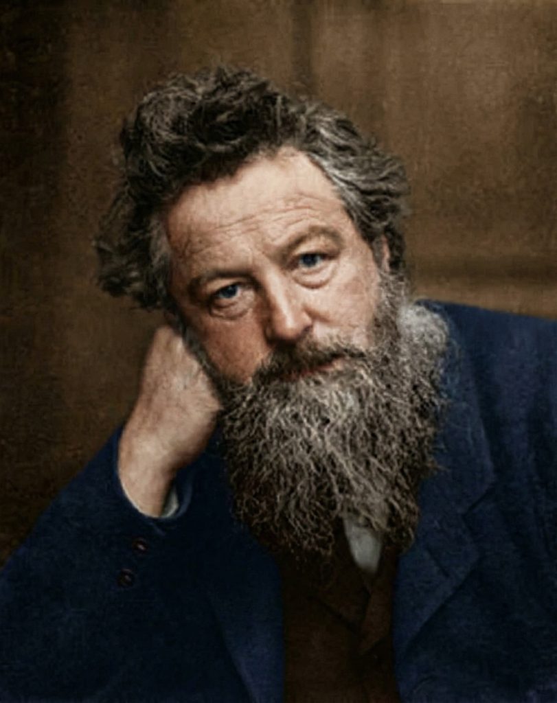 Colorized portrait of William Morris, founder of the Arts and Crafts movement, resting his head on his hand, with a full grey-streaked beard and intense blue eyes, circa 1880s