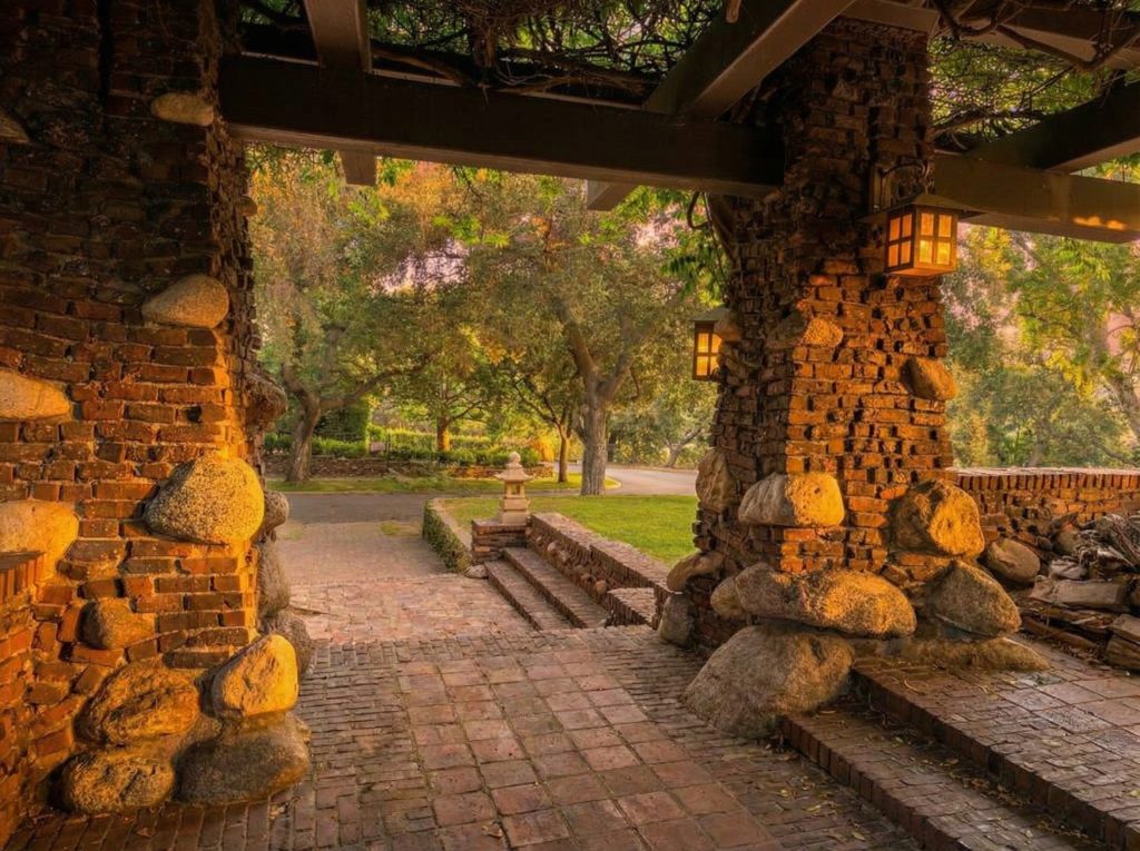 Photograph looking out from beneath the Duncan-Irwin House pergola at golden hour, framed between two massive clinker-brick-and-river-stone piers with hanging copper lanterns, looking past brick paving toward a Japanese stone lantern on the lawn and mature oak trees in the garden beyond