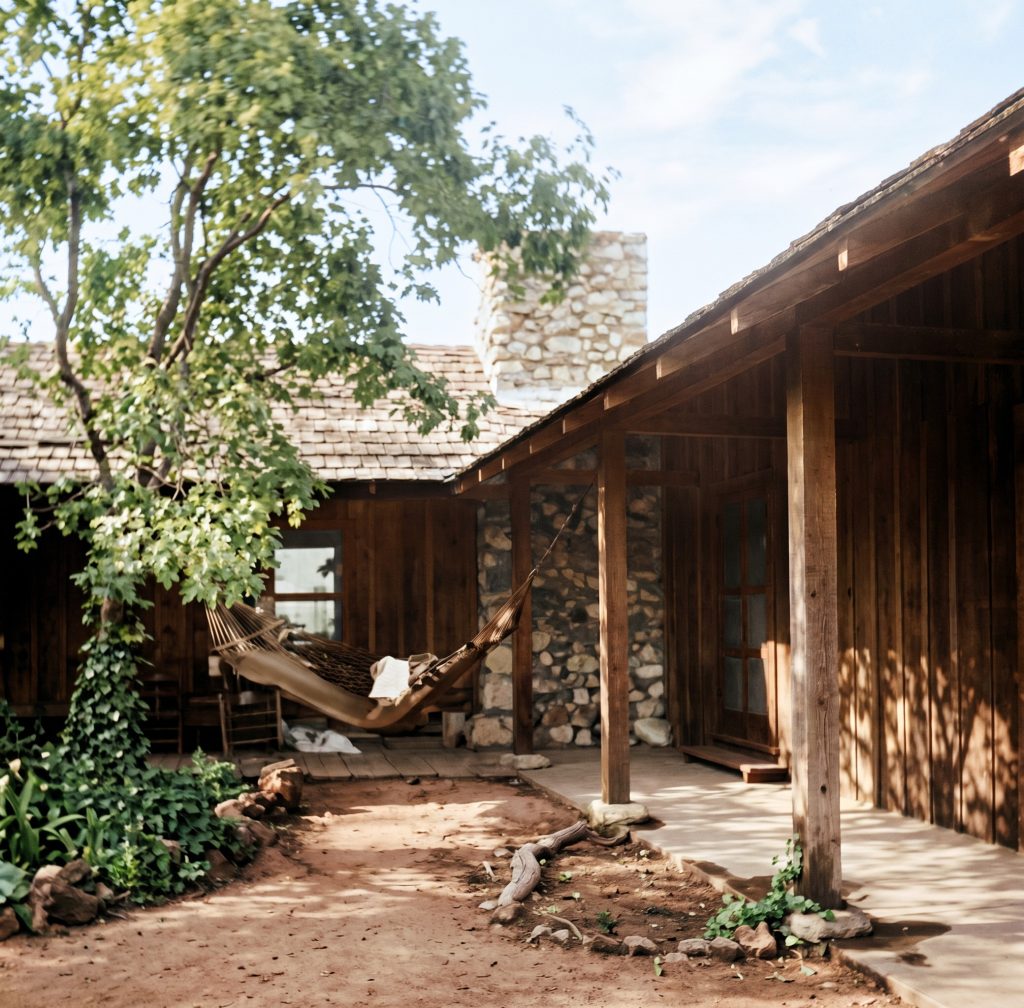 A man reclines in a hammock on a covered porch beside an open, sunlit courtyard; historic photograph, colorized.