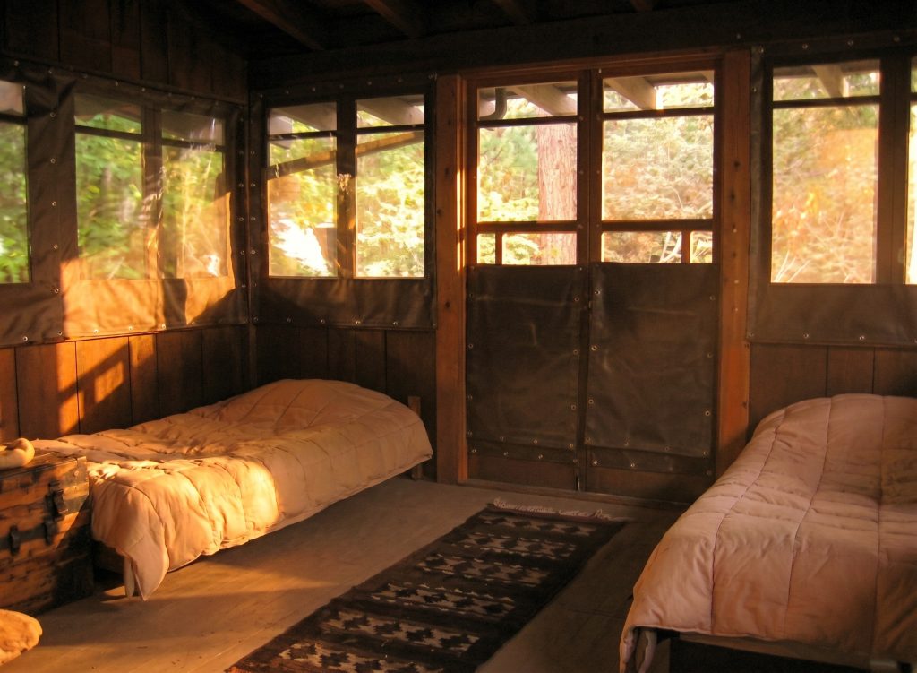 Interior view of one of the three sleeping porches at the Pratt House, a screened upstairs room opening to the valley view with a low bed and the wood ceiling above.