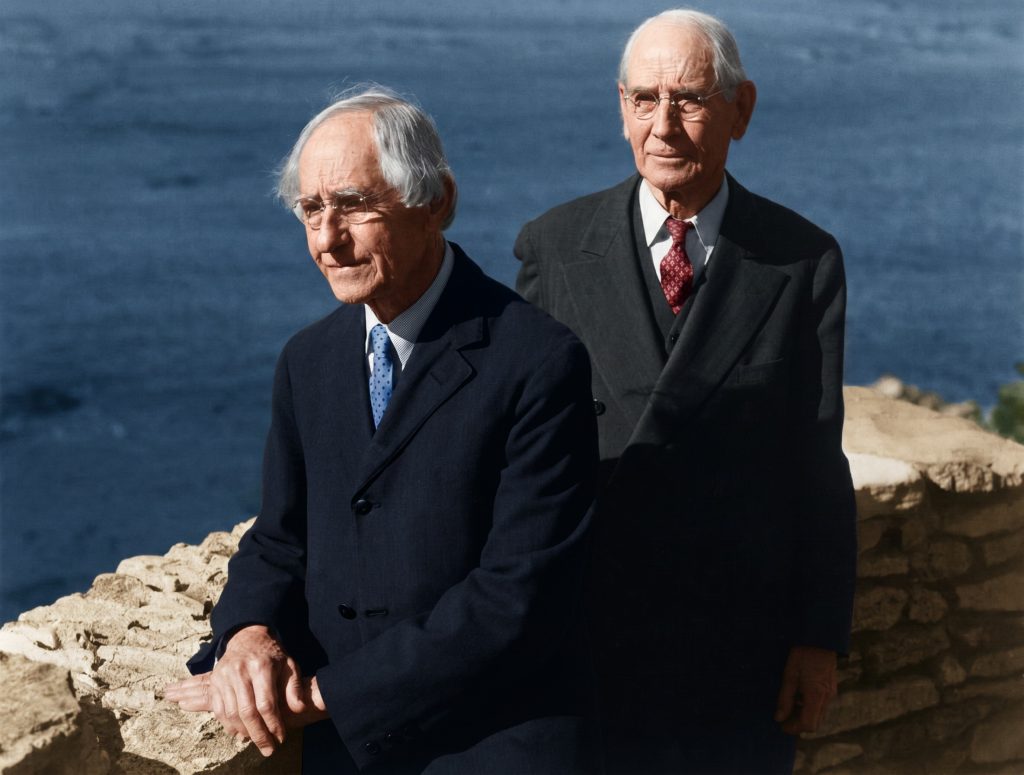 Color photograph of the elderly Greene brothers standing together in front of a low stone wall with the Pacific Ocean visible behind them, Charles Sumner Greene at left wearing a navy overcoat and blue tie, Henry Mather Greene at right wearing a dark suit and red patterned tie, both with white hair and the bearing of men in their eighties