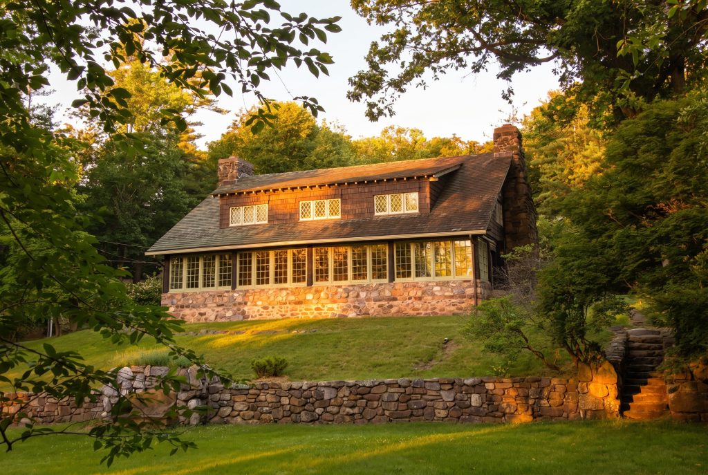Exterior view at golden hour of Craftsman Farms, Gustav Stickley's home in Parsippany, New Jersey, a long low building with a stone foundation, brown shingle siding, deep eaves, a row of casement windows with diamond-paned upper sashes, a stone chimney, and a coursed-stone retaining wall in the foreground beneath a lawn.