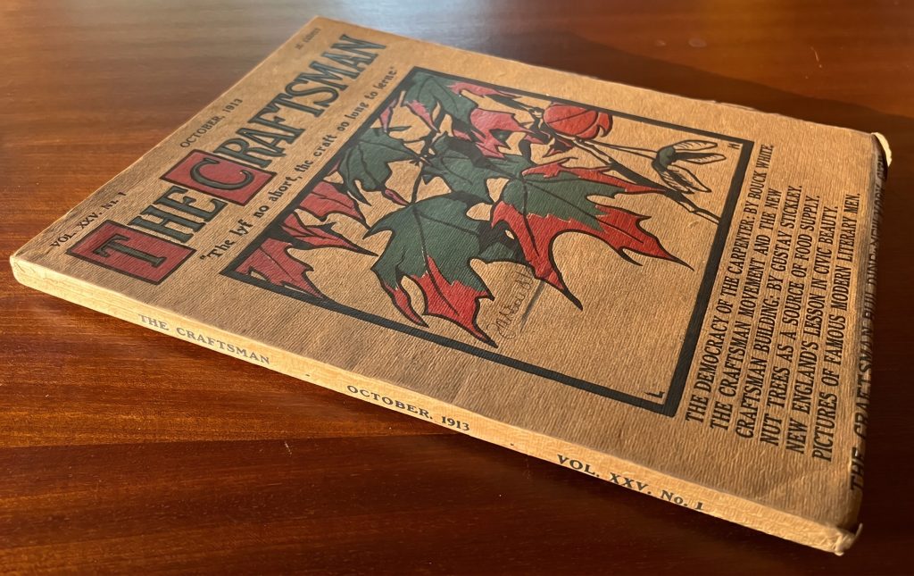 A bound issue of The Craftsman magazine, Volume XXV, Number 1, dated October 1913, photographed at an angle on a wood surface, with its cover showing a stylized illustration of red and green leaves on a tan ground, the title in red letters at upper left, and a list of feature articles printed vertically along the right margin including "The Democracy of the Carpenter," "The Craftsman Movement and the New Craftsman Building," and "Nut Trees as a Source of Food Supply."