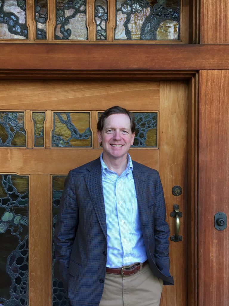 Edward R. Bosley standing in front of the Gamble House front entry, smiling at the camera. He wears a checked gray sport coat over a striped blue dress shirt and tan trousers. Behind him, the Tiffany-style leaded art glass of the front door depicts a tree-of-life motif in amber, green, and bronze tones within mahogany framing; a hammered copper door pull is visible at right.