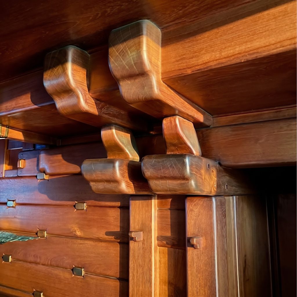 Close-up detail of the carved cloud-lift outrigger brackets at the upper level of the Gamble House staircase, showing the deeply rounded shoulder profiles in teak with visible grain patterns, photographed in strong raking afternoon light that emphasizes the three-dimensional carved form, with rows of exposed strap hardware fastening the horizontal-board wainscot panels below.