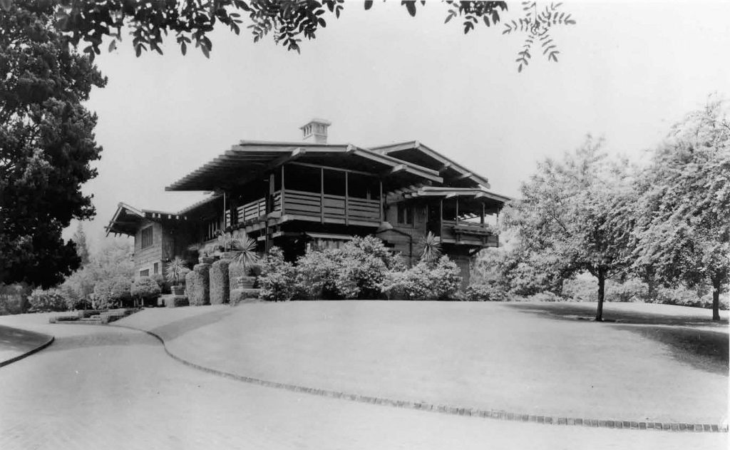 Black-and-white photograph of the Gamble House from a side-driveway view in 1956. The two-story shingled Craftsman residence is visible with its broad overhanging eaves, second-story sleeping porches, central rooftop cupola, and surrounding mature plantings. A curving paved drive sweeps across the foreground.