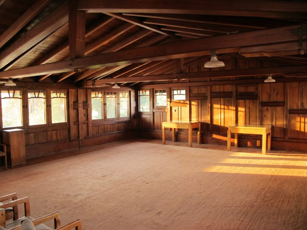 Wide view of the Gamble House attic looking the length of the room toward a gable end, showing the exposed rafter ceiling, paneled walls, a band of windows with art-glass transoms along both eaves, and late-afternoon sunlight raking across the wood floor.