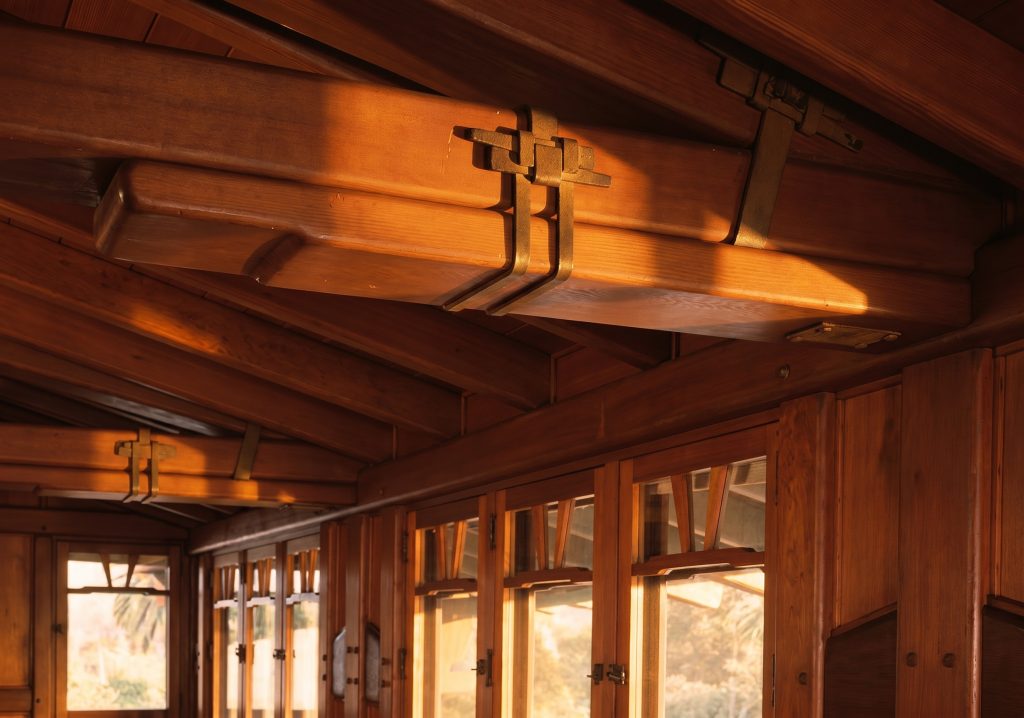 Close-up of a flat iron strap painted soft green-black wrapping the underside of the Gamble House attic ridge beam, riveted and bolted into the timber with hexagonal bolt heads and domed rivets fully visible, the warm grain of the surrounding fir framing lit by horizontal afternoon sun.