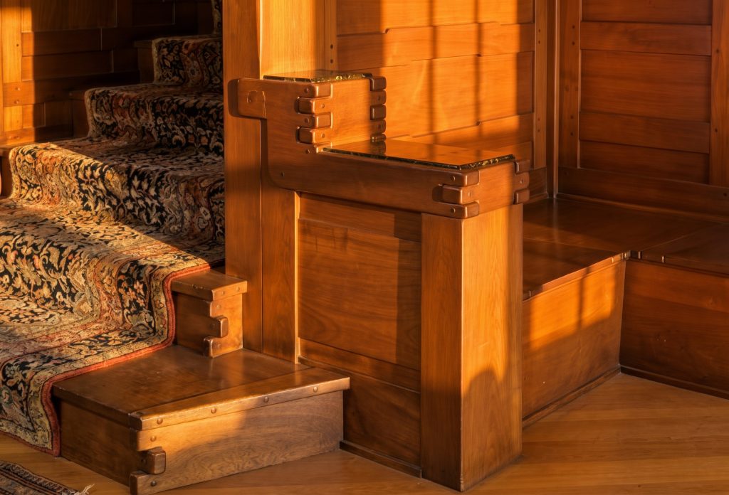 Close view of the Gamble House staircase newel post and built-in bench in warm morning light, showing the stacked horizontal bands of the newel cap with visible fasteners and a small marble set flush into the top, the wide teak bench seat, an outer step platform at the base of the newel, and the dark floral carpet runner on the stair treads at left.