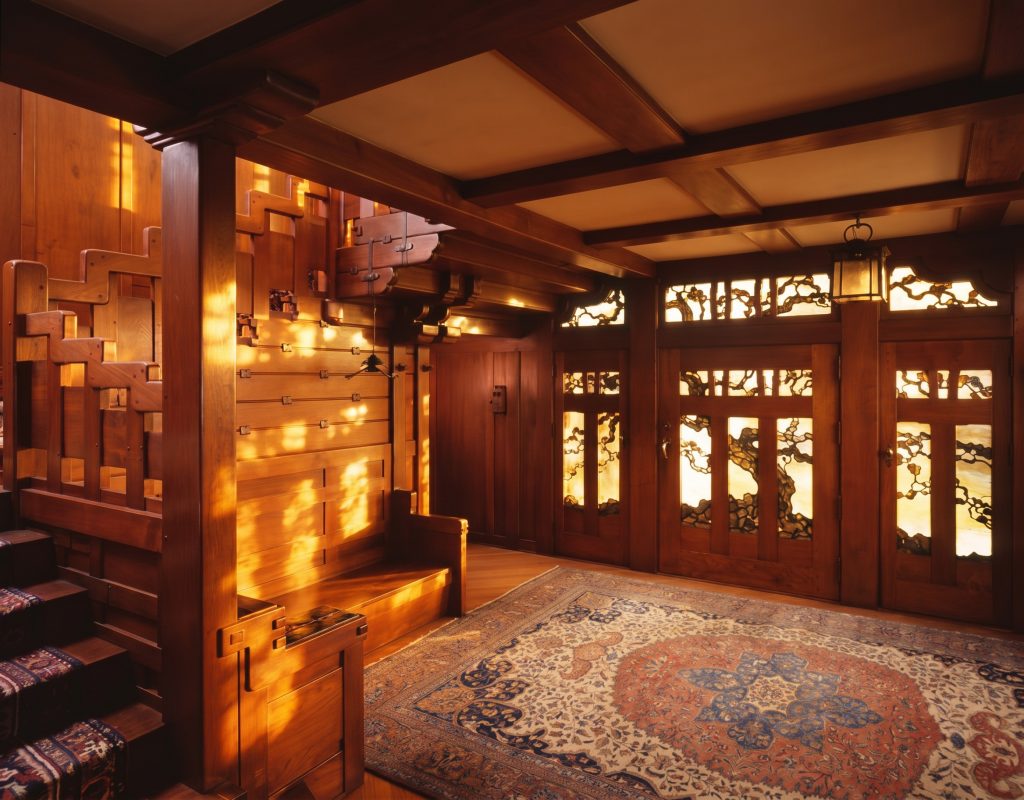 Wide view of the Gamble House entrance hall showing the staircase on the left with stepped cloud-lift balustrade and built-in bench at the foot, the coffered ceiling with rice-paper panels overhead, a hanging period lantern near the front door, Charles Greene's Japanese black pine art-glass front door and sidelight panels glowing amber and gold at right, and a large Persian rug on the entry hall floor.