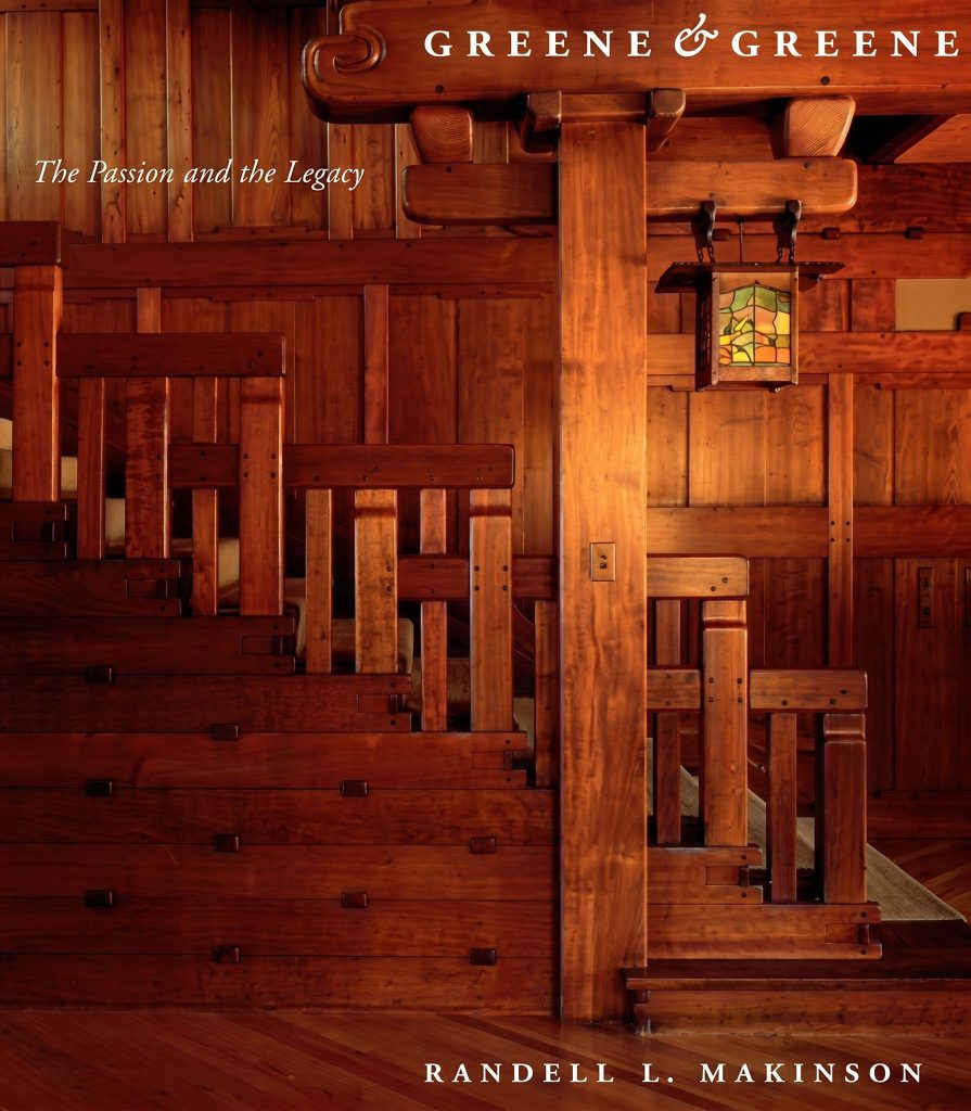 Cover of Randell L. Makinson's 1998 book Greene & Greene: The Passion and the Legacy. A color photograph of a Greene & Greene mahogany staircase fills the cover, showing stepped tread caps, square ebony pegs, paneled stair-side construction, a carved scroll bracket at top, and a leaded art glass lantern hung from a ceiling beam. Title in italic serif type at upper left; Makinson's name at lower right.