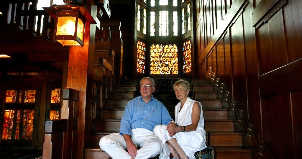 Harvey and Ellen Knell, restorers of the Blacker House, seated together on a Greene & Greene staircase landing. Harvey wears a light blue button-down shirt and white trousers; Ellen wears a white sleeveless top. Behind them, a backlit leaded art glass window glows with intricate jewel-toned patterns. A copper-and-glass lantern hangs at left; mahogany paneling, balustrade, and stair construction fill the frame.