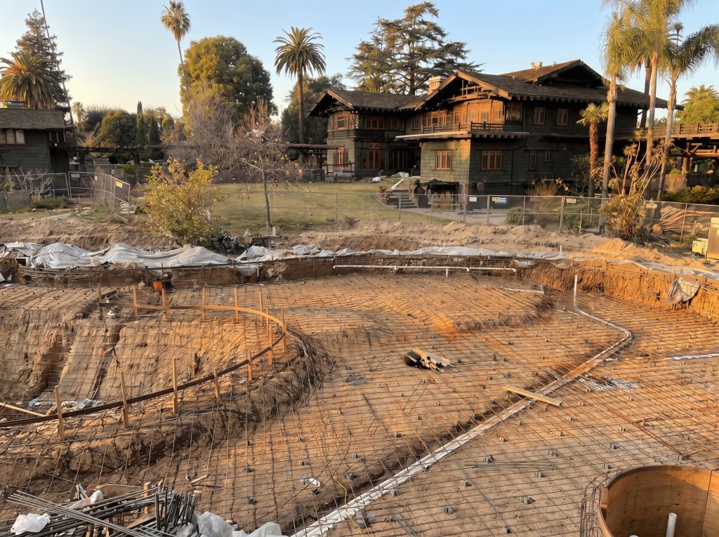Contemporary photograph of pond reconstruction in progress in the rear garden of the Blacker House, showing a large biomorphic excavated basin with steel reinforcing mesh laid across the floor, temporary chain-link fencing separating the work zone from the lawn, and the Blacker House standing intact behind palms and pergolas in the background.