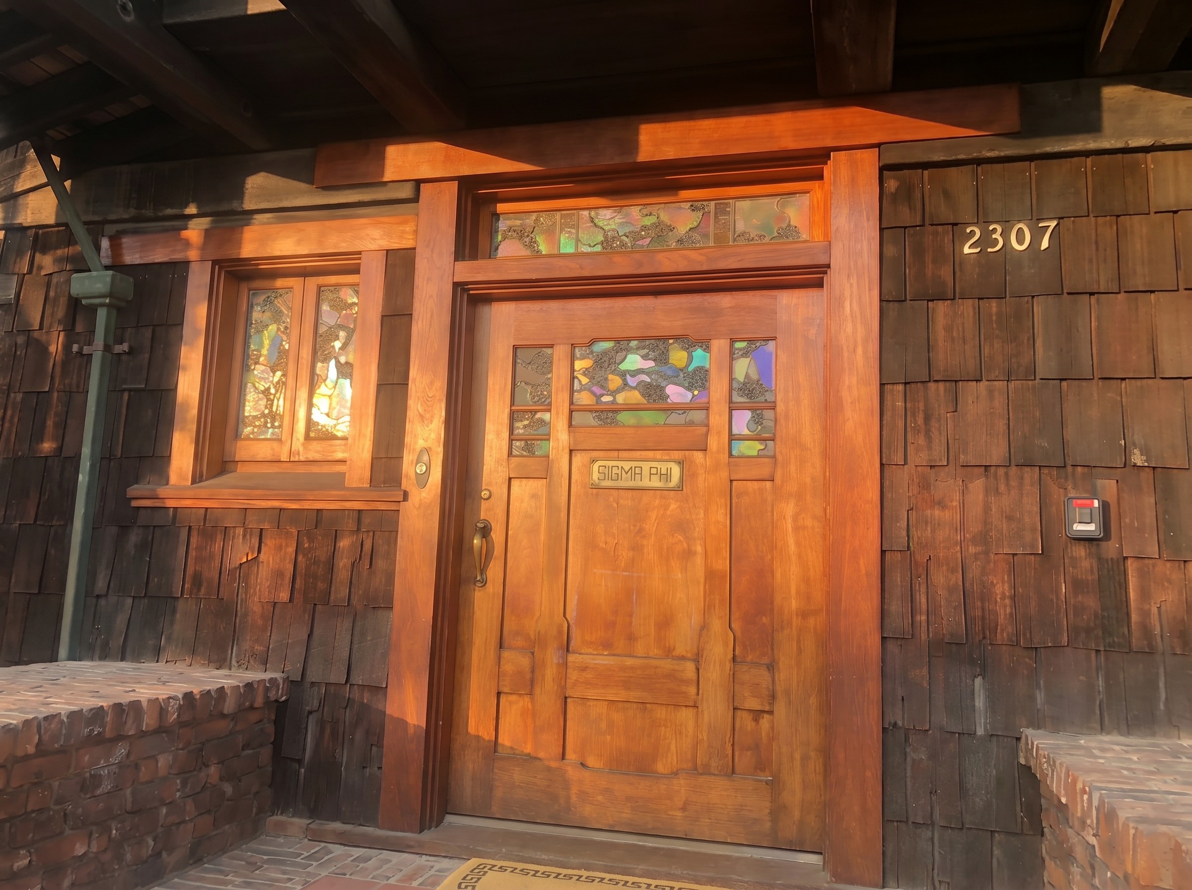The front door of the Thorsen House from outside, a broad figured-wood door with a band of leaded art glass across the top, a brass SIGMA PHI plate, and the house number 2307 on the shingled wall.