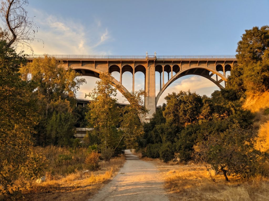 The Colorado Street Bridge in Pasadena seen from a dirt path on the floor of the Arroyo Seco at sunset, its tall concrete arches rising above oak and sycamore canopy with the canyon's dry grasses in the foreground.