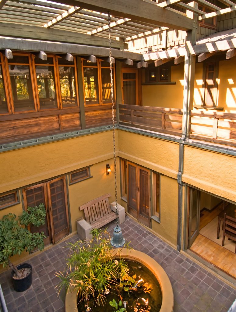Overhead view of the Duncan-Irwin House courtyard from the second-floor balcony, showing the circular lily pond, brick paving, stucco walls of the lower story, and shingled second-story walls beneath an open pergola.
