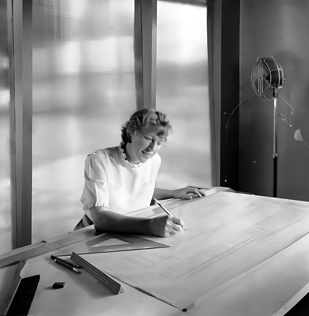 Esther McCoy seated at a drafting table, leaning over a sheet of architectural drawings with a pencil in her right hand. She wears a short-sleeved white blouse. A T-square, parallel rule, and pencils rest on the drafting surface; a stand fan stands at right against tall windows that wash the room in soft light.
