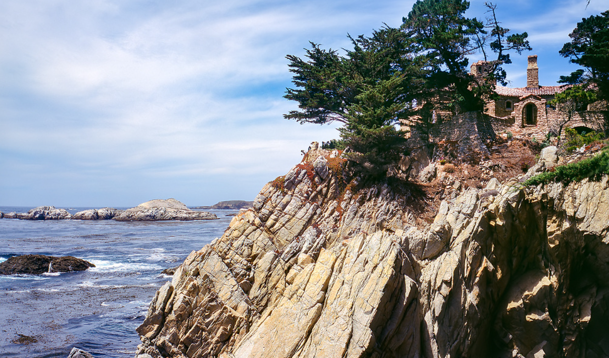 Distant view of D.L. James House perched on granite cliffs above the Pacific Ocean at Yankee Point in Carmel Highlands, California, with layered stonework rising directly from the bedrock, terra-cotta roofs, multiple terraces, and Monterey cypress trees framing the composition