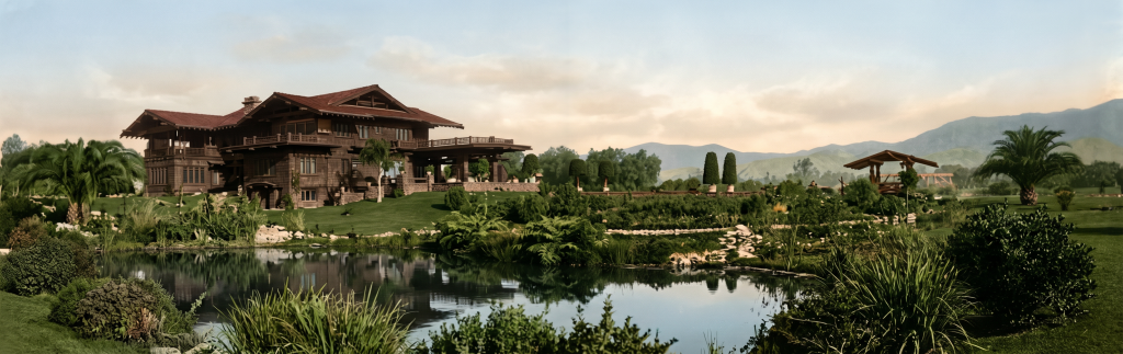 Colorized panoramic historic photograph of the Robert R. Blacker House in Pasadena seen across the original lily pond, with the house elevated on its clinker brick podium at left, the pond reflecting the sky in the foreground, palms and tropical plantings framing the view, and the San Gabriel mountains rising in the distance.