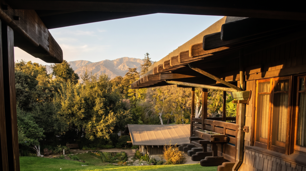 View from a second-floor sleeping porch at the Gamble House, looking past the deeply overhanging timber eaves toward the San Gabriel mountains in the distance, with the garden and a lower porch roof visible below.