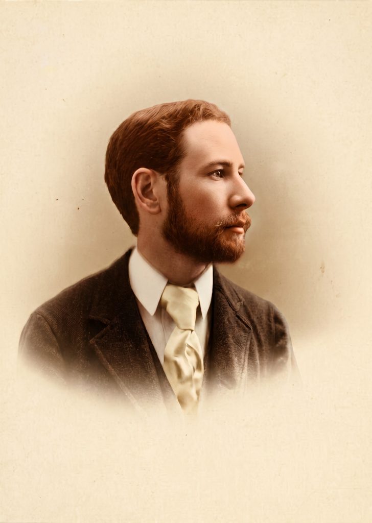 Colorized historic studio portrait of Henry Mather Greene shown in three-quarter profile, with reddish-brown hair and full reddish beard, wearing a dark wool jacket, white wing-collared shirt, and pale silk cravat, photographed against a soft sepia studio backdrop