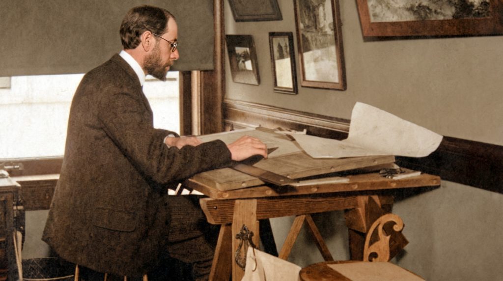 Colorized historic photograph of Henry Mather Greene seated at a wooden drafting table, shown in profile wearing a tweed jacket, white collar, and round glasses, with reddish-brown hair and beard, his hand resting on a set of architectural drawings spread across the table, with framed renderings hung on the wall behind him and a window casting natural light across the workspace