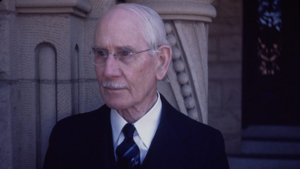 Color photograph of Henry Mather Greene late in life, shown in profile in his eighties, with white hair and a white mustache, wearing wire-rimmed glasses, a dark suit, white shirt, and striped navy tie, standing in front of a carved stone architectural element