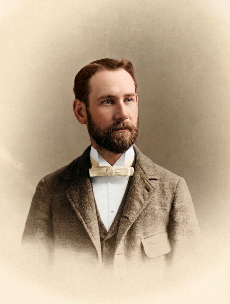 Colorized historic studio portrait of Henry Mather Greene shown in three-quarter view facing left, with brown hair and a full brown beard, wearing a tweed three-piece suit with white wing-collared shirt and pale bow tie, photographed against a soft sepia studio backdrop