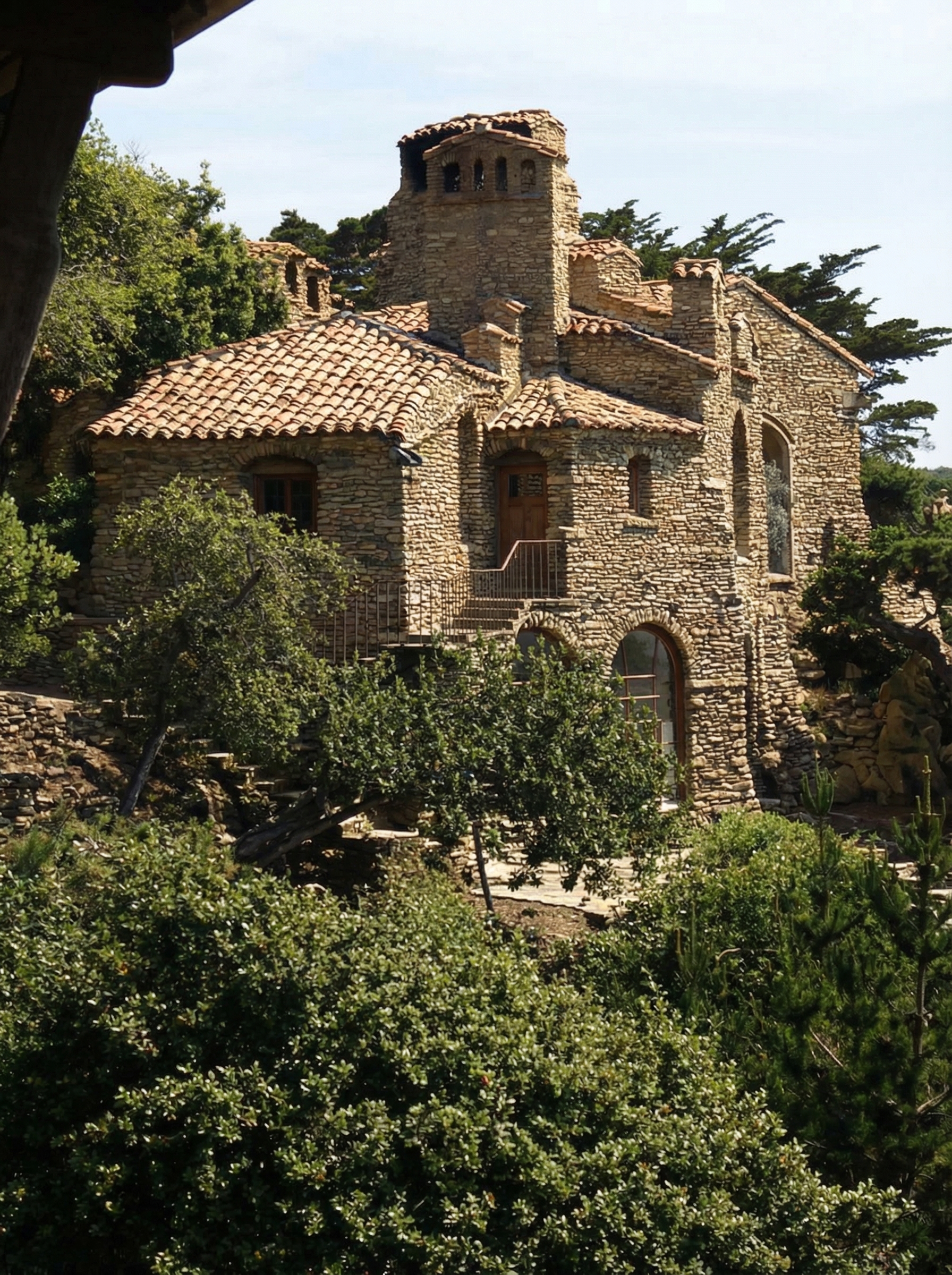 Historic colorized photograph of the D.L. James House from the west late in the afternoon, showing the stepped terraces descending toward the Pacific Ocean, the granite walls glowing in low western sunlight, the terra-cotta roofs catching the same warm light