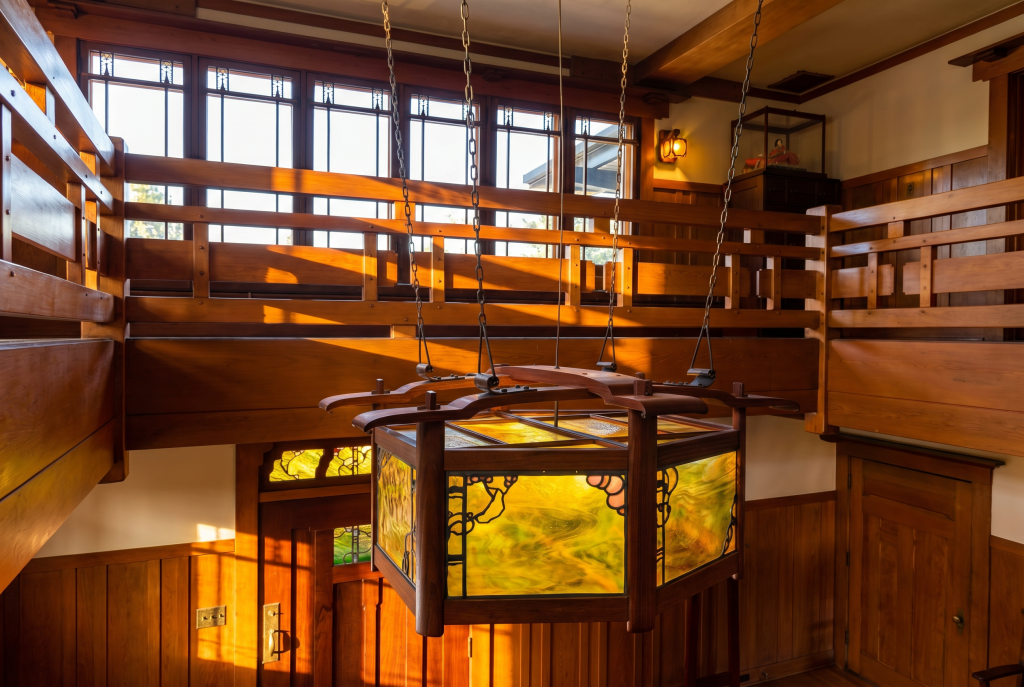 Photograph of the same Robinson House lantern read from the second-floor landing, showing the mahogany frame with curved upper rim, the amber and green leaded art-glass panels with lead-work in floral and curvilinear motifs, the iron chain suspensions to the ceiling, and the clerestory windows of the upper hall in the background