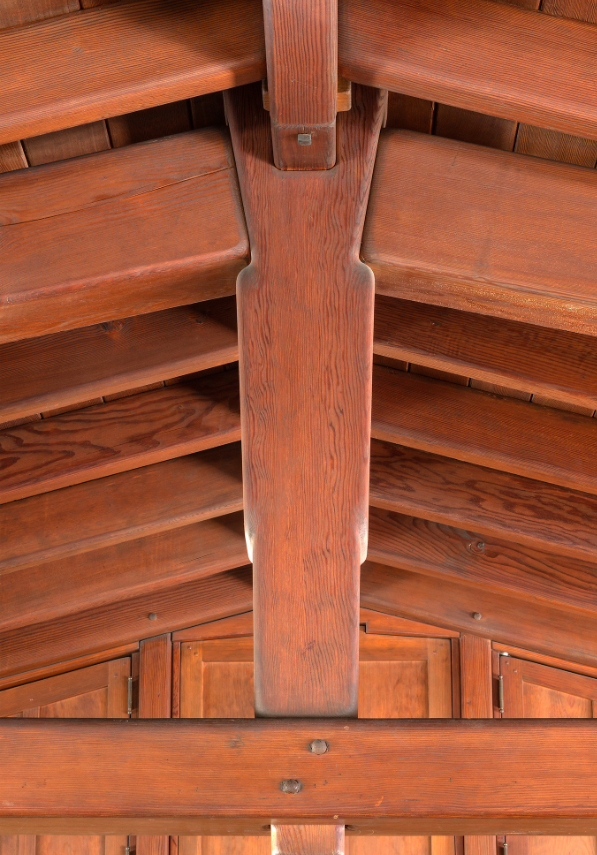 Close-up looking up at a king-post detail in the Gamble House attic, showing a vertical wood member meeting a horizontal tie beam with an ebony peg at the joint, flanked by symmetrical pairs of rafters and the diagonal lines of the boarded ceiling.