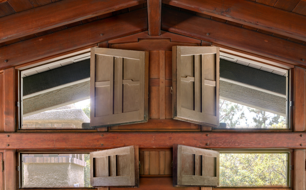 The Gamble House attic gable shutters shown in the opened position, swung outward to reveal the diagonal art-glass-trimmed windows behind them admitting bright daylight, with the framed gable peak above and the exposed rafter ceiling rising overhead.