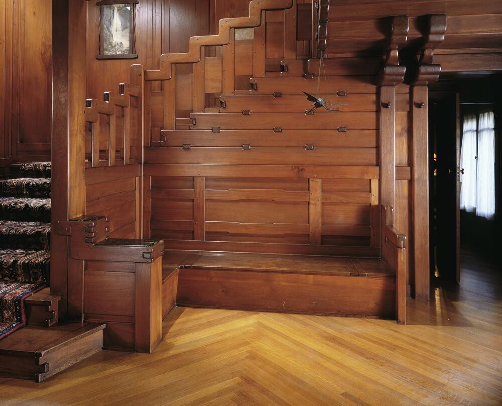 Close view at the foot of the David B. Gamble House staircase showing a built-in landing bench, the stepped paneled wainscot above with exposed metal strap-and-bolt joinery fastening the panels to the structure beneath, a paisley runner climbing the treads at left, a bronze crane figure suspended on a thin line at center, herringbone oak flooring, and a corridor opening at right.