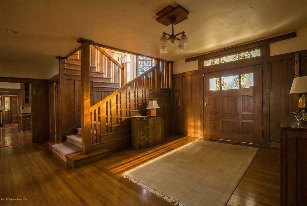 Wider view of the William T. Bolton House entry hall showing the staircase rising on the left with carpet runner and square balusters, a low cabinet with painted decoration beneath the stair, a large front door at right with leaded-glass panels in a daffodil pattern above and at the side, a multi-bulb period chandelier on a wood ceiling block, and polished wood floors with a runner.