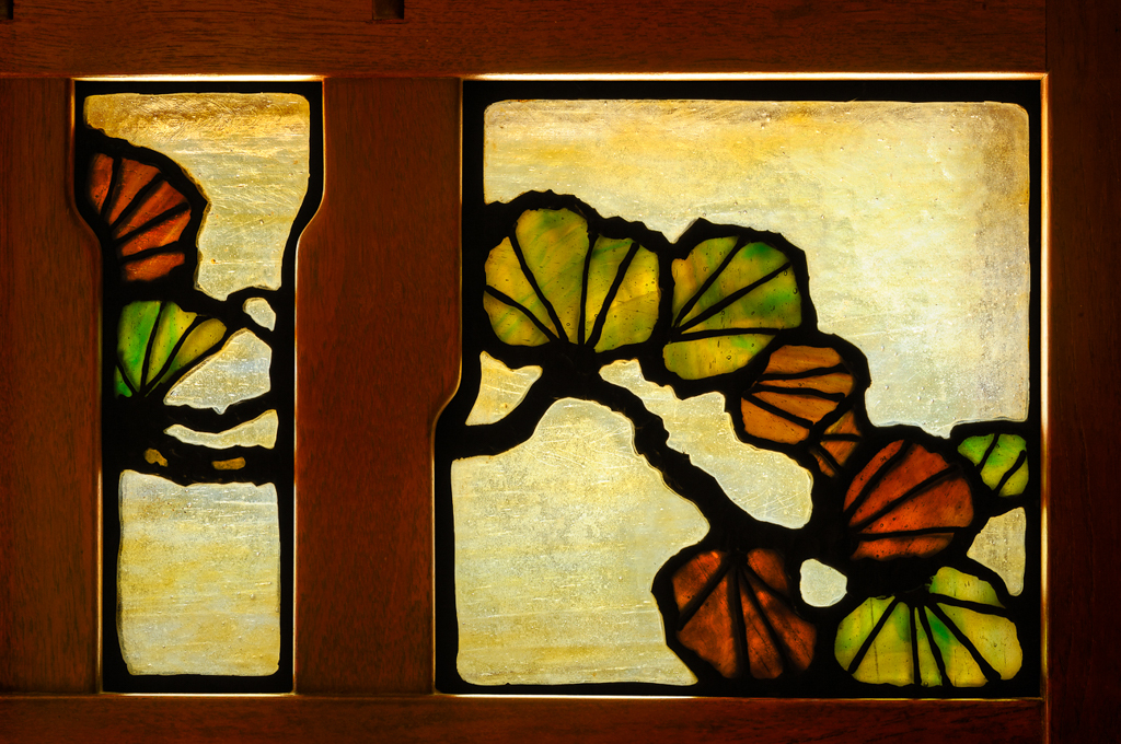 Close-up detail of the front door art glass at the David B. Gamble House in Pasadena, showing the leaded art-glass triptych of central door and two sidelights with a single Japanese black pine spreading its branches across all three panels, the iridescent rolled glass and copper-foil leading visible against the dark teak frame
