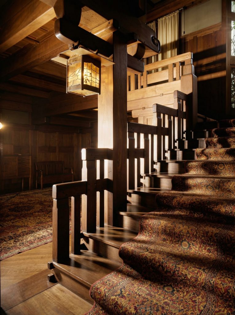 Close view of the famous Robert R. Blacker House staircase newel post in deep mahogany, with a deeply carved scroll capping the cap-rail, a square mica-and-leaded-glass lantern hanging from the scroll on a chain, paneled balustrade with peg detail climbing to the right, paisley Persian runner on the carpeted treads, wood plank floor and oriental rug at left, and drapery beyond a window at upper right.