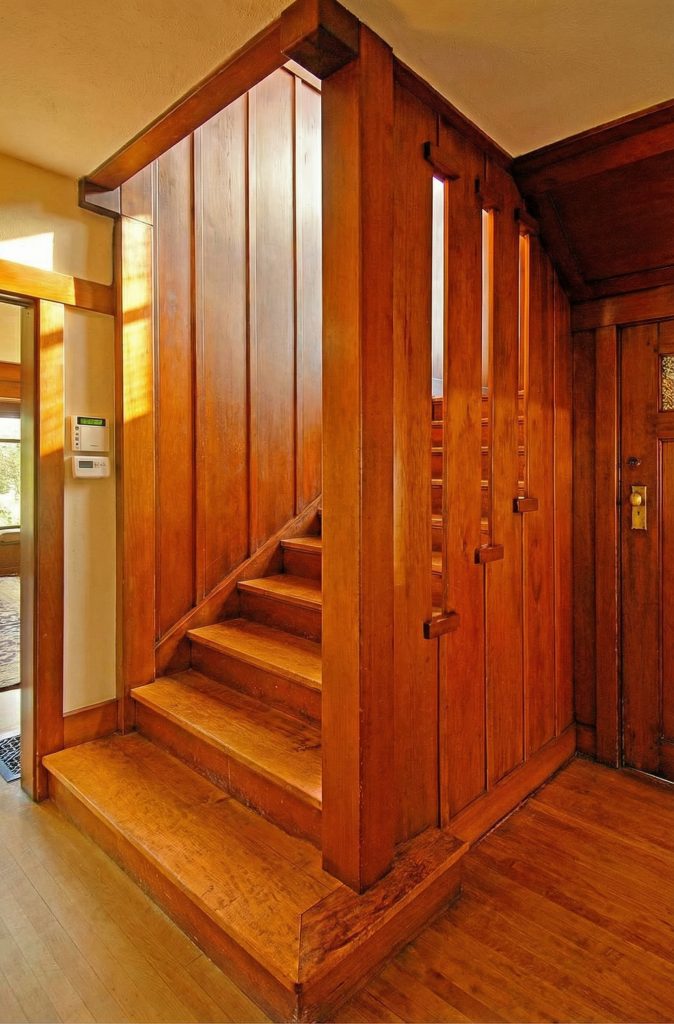 View of the Caroline S. de Forest House staircase showing a narrow vertical-board paneled stair tower with three slim vertical slot openings admitting light at the upper level, a sliding pocket door at right with a brass knob, a glimpse of the front door area with a rug at left, and polished wood floors.