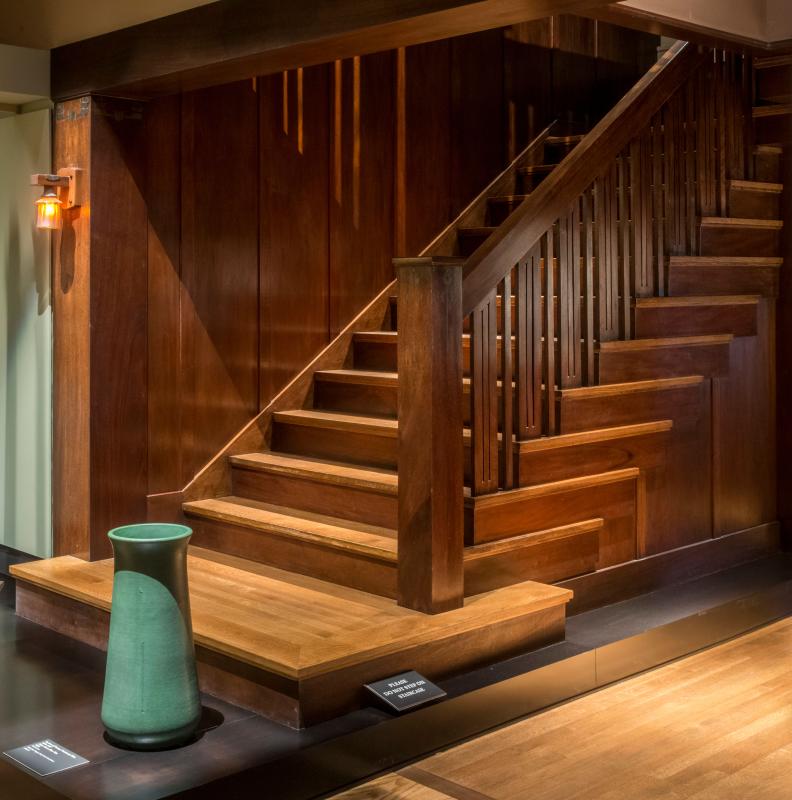Museum installation at the Huntington Library showing the Dr. Arthur A. Libby House staircase with stepped paneled wainscot, slim square balusters, a square newel, a copper carpenter's lantern wall sconce at upper left, a low platform with a green vase displayed in front of the stair, a "Please do not touch" sign on the platform, and gallery flooring.