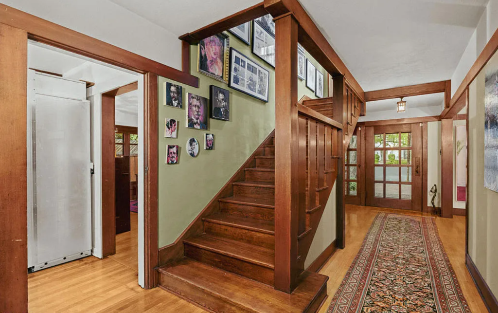 Late-afternoon view of the Louise C. Bentz House staircase with a sloped-rail balustrade descending into a sitting area, a mica-shaded table lamp on a low wood chest at left, an open garden door at right admitting golden light, a woven rug on the raised platform, drapery, and a vase of bare branches in the corner beyond.