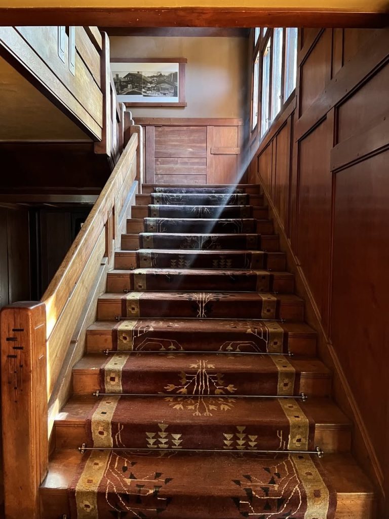 Looking up the William R. Thorsen House staircase from below, with a stylized period runner secured to each riser by brass stair rods, a heavy beam of late-afternoon sunlight slanting in from a high window on the right, paneled wainscot with exposed peg details climbing to the left of the runner, a small framed photograph hung at the landing, and dark wood paneling throughout.