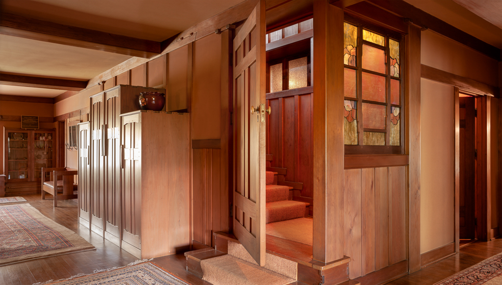 The second-floor hall of the Gamble House looking toward the open door of the attic stair, with an art-glass panel of pale ginkgo leaves set into the door, a flight of carpeted stairs rising behind it, and built-in storage cabinets along the hall wall.