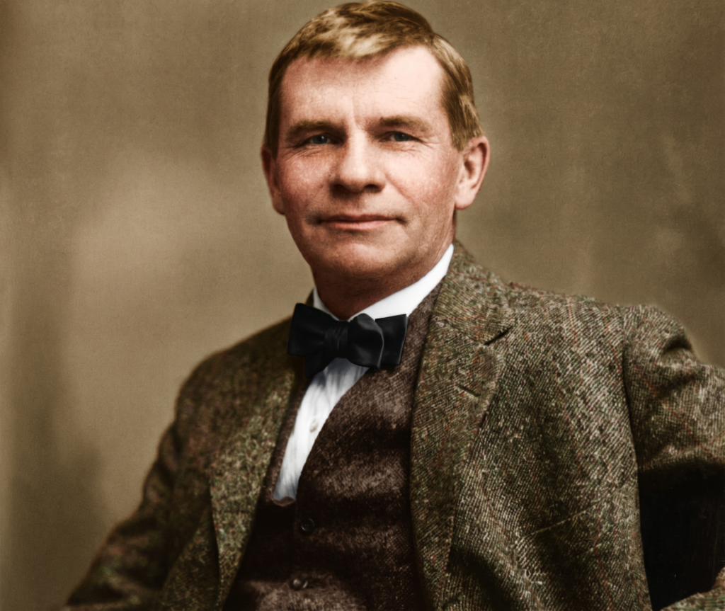 Color-tinted formal portrait of Gustav Stickley, a fair-haired man in his middle years wearing a flecked tweed three-piece suit and a black bow tie, photographed seated against a plain studio backdrop, looking to the side of the camera with a slight smile.