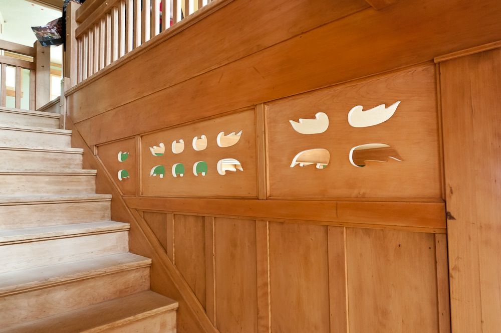 Close view of the Jennie A. Reeve House staircase wainscot panels showing pierced cutout shapes in stylized geometric and folk-art forms — abstracted birds, fish, leaves, and curving motifs — set into flat painted panels backed with green and amber color, with the stair treads rising at left and additional vertical-board paneling below.